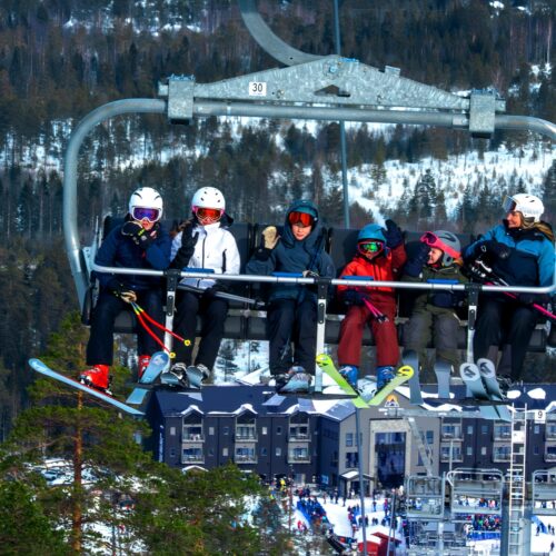 Seks personer sidder i en skilift med ski, snedækkede træer og bygninger i baggrunden.