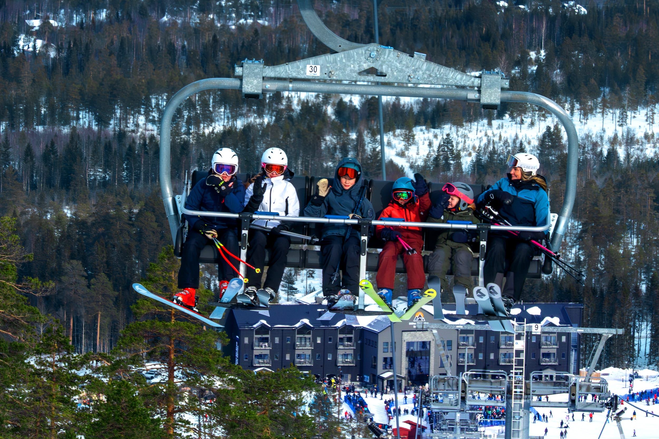 Sex personer sitter i en skidlift med skidor, snöklädda träd och byggnader i bakgrunden