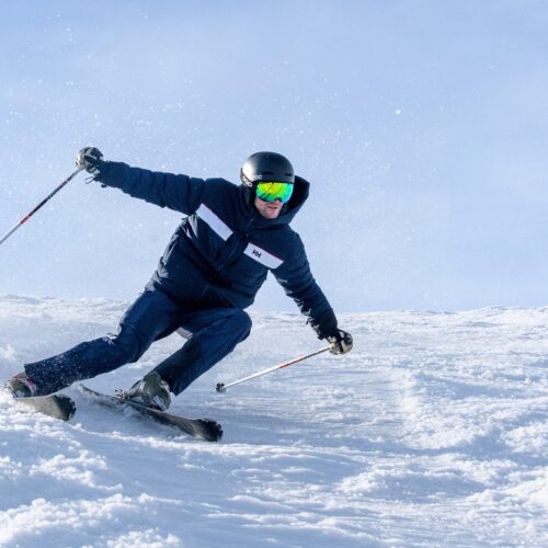 Person i mørk skijakke står på ski ned ad snedækket skråning med skistave, sneen hvirvler op mod lyseblå himmel
