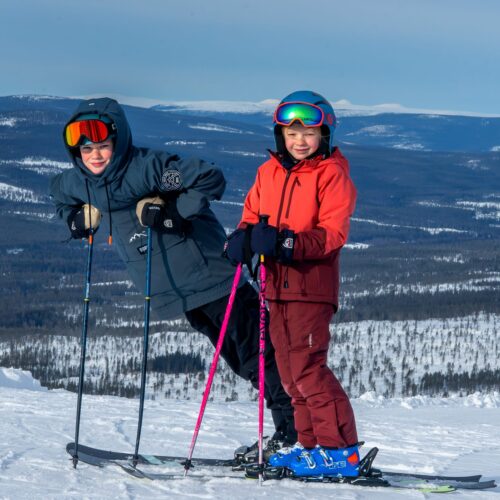 Två personer i skidkläder och hjälmar står med skidor och stavar på snötäckt berg med utsikt över fjällandskap