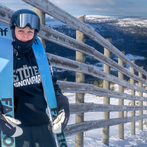 Person i sort sweater holder blå ski ved træhegn med snedækket bjerg i baggrunden