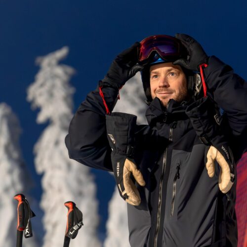 Person i mörk skidjacka justerar skidglasögon, snötäckta träd och röd säkerhetsnät i bakgrunden