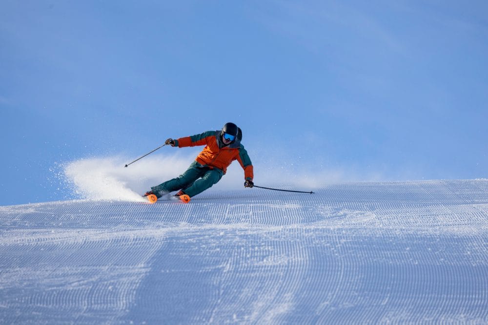 Person in orange ski jacket skiing down freshly groomed snow-covered slope with ski poles under blue sky