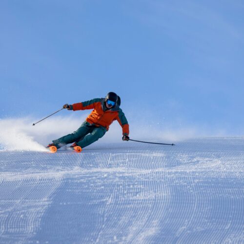 Person i orange skijakke står på ski ned ad nypræpareret snedækket piste med skistave under blå himmel
