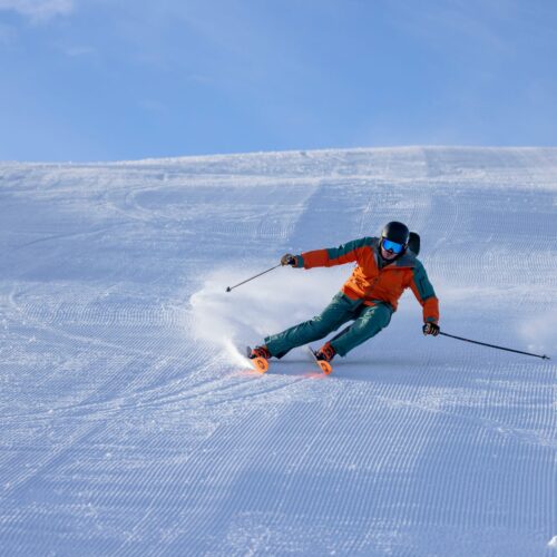 Person i orange skijakke står på ski ned ad nypræpareret snedækket piste med skistave under blå himmel