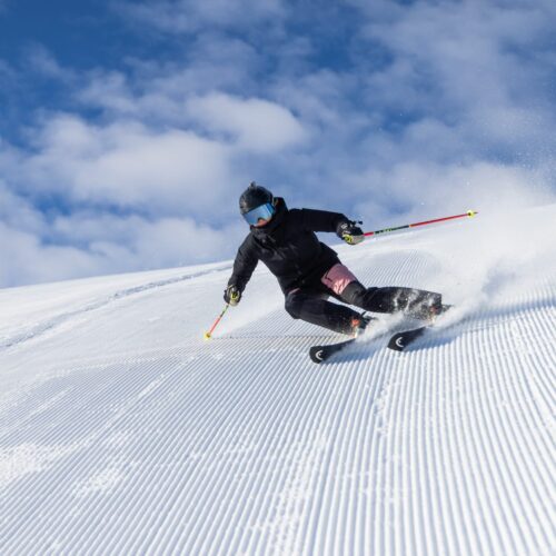 Person åker skidor nedför bred snötäckt backe med skidstavar, snön virvlar upp mot blå himmel