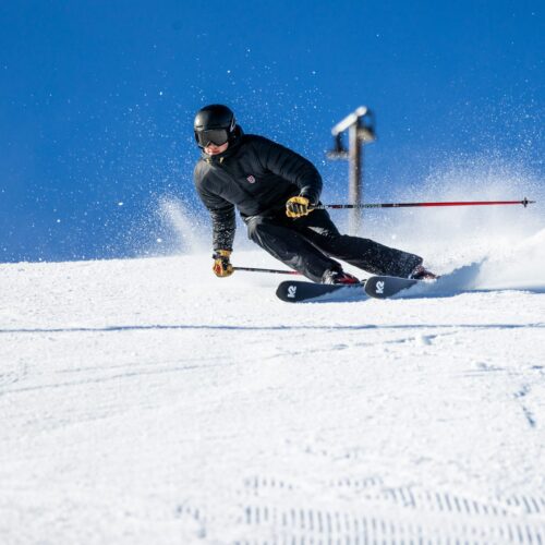 Person in black ski jacket skiing down snow-covered slope with ski poles, snow spraying up against clear blue sky