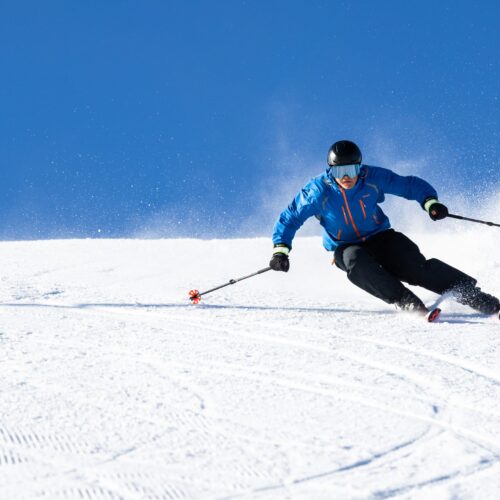 Person in blue ski jacket skiing down snow-covered slope with ski poles, snow swirls up against clear blue sky