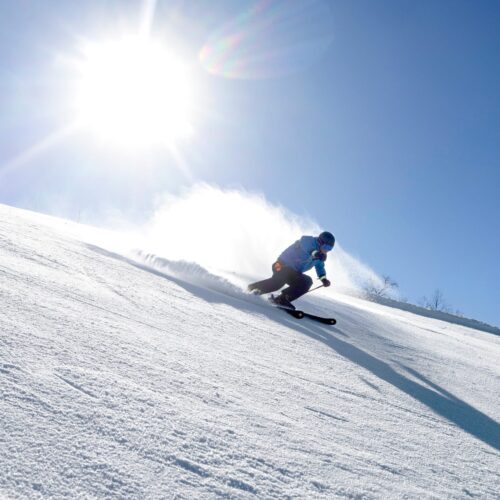 Person i blå skidjacka åker skidor nedför snötäckt backe, snön virvlar upp mot stark sol och blå himmel