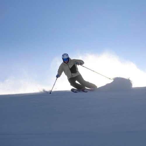 Person åker skidor nedför snötäckt backe med skidstavar, snön virvlar upp mot klar blå himmel