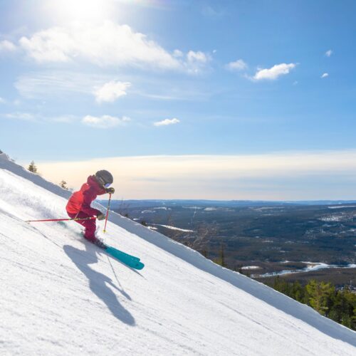 Person i rød skijakke står på ski ned ad snedækket skråning med blå ski under solrig himmel