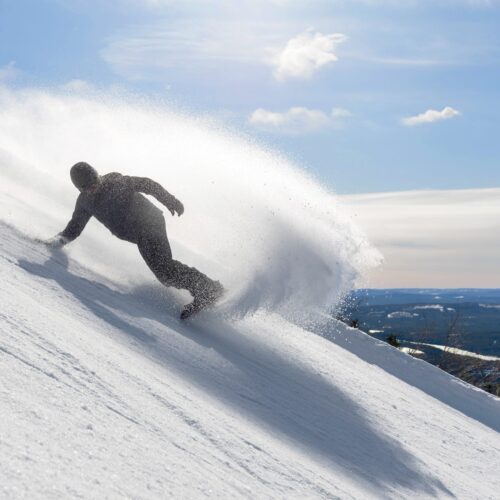 Person åker snowboard nedför snötäckt backe, snön sprutar upp mot klar blå himmel