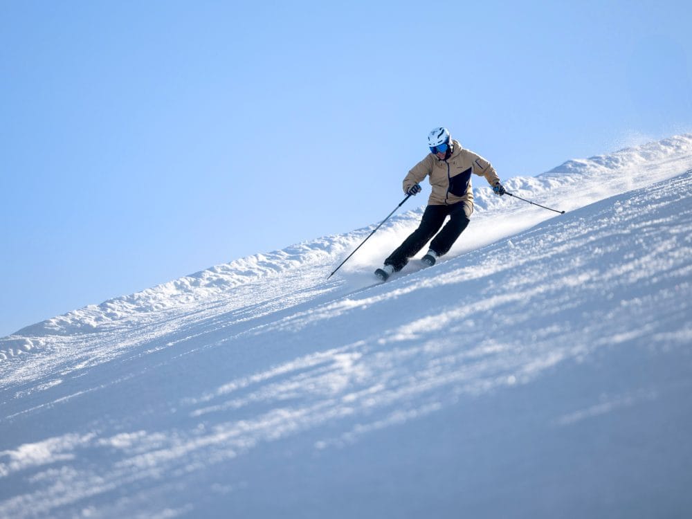 Person på ski ned ad snedækket skråning med skistave under klar blå himmel