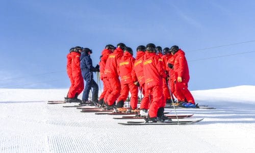 Group of ski instructors in red jackets and helmets standing on snow-covered slope under blue sky