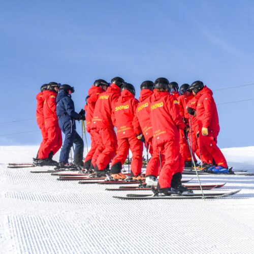 En gruppe skiinstruktører i røde jakker og hjelme står på en snedækket skråning under blå himmel.