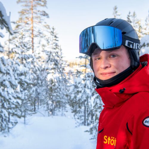 Person i rød skijakke med hjelm står blandt snedækkede træer i vinterlandskab