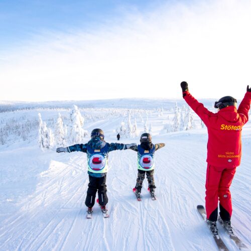 Instruktör och två barn i skidskola står med utsträckta armar på snötäckt fjälltopp