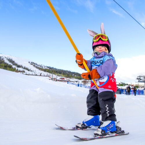 Barn i skidskola åker lift med hjälm och skidutrustning på snötäckt skidbacke