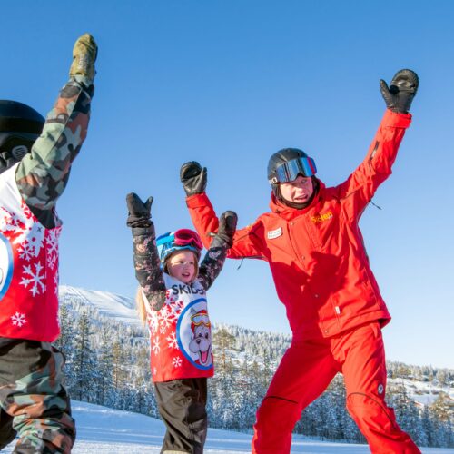 Instruktør og børn i skiskole står med løftede arme på solbeskinnet skibakke