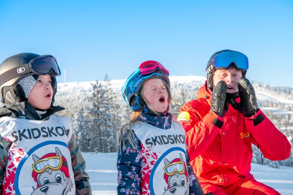Två barn i skidskola med instruktör i röd jacka på solig skidbacke