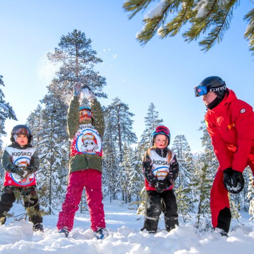 Barn i skidskola leker i snön med instruktör bland snötäckta träd i solsken