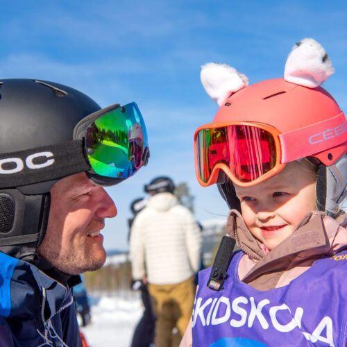 Två barn i skidutrustning står nära varandra under skidskola på solig skidort