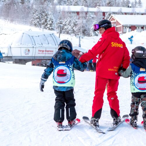 Instruktør hjælper to børn i skiskole ved liftområde i snedækket skisportssted