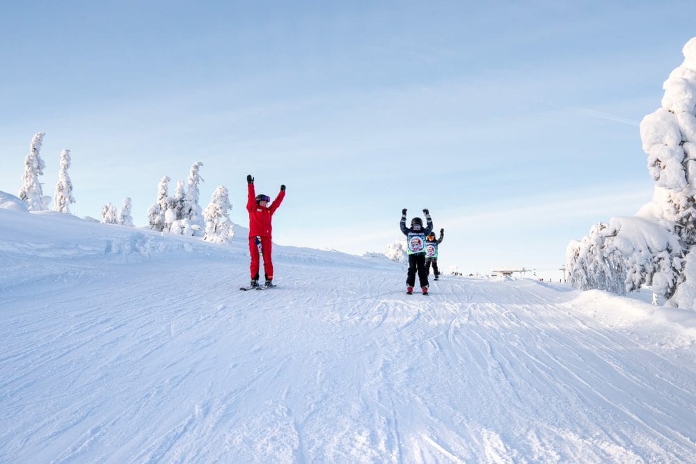 Instruktör och barn i skidskola åker i solig backe med snötäckta träd omkring