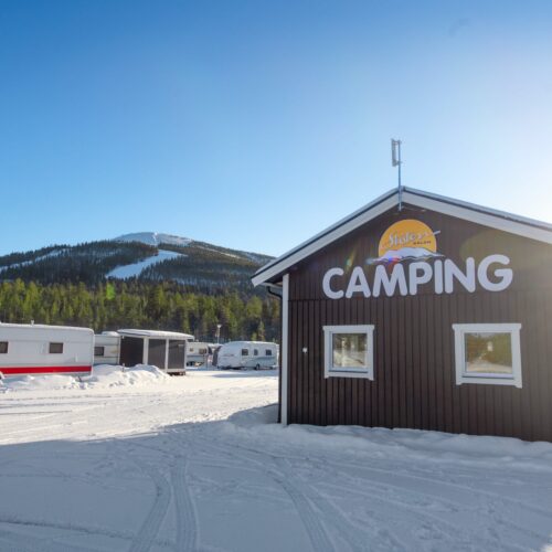 Brun bygning med teksten “Camping” på facaden, snedækket jord, campingvogne og bjerge med en skibakke i baggrunden under en klar blå himmel.