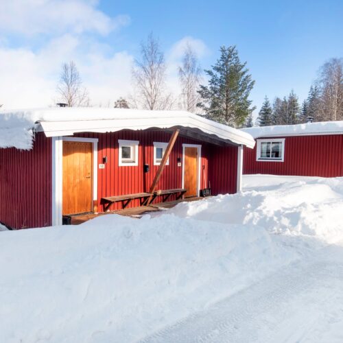 Röd stuga med vita knutar och snötäckt tak omgiven av vinterlandskap.