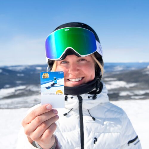 Person in white ski jacket holding up ski pass with snow-covered mountains in the background