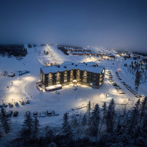 Illuminated mountain hotel surrounded by snow-covered ski slopes and parked cars on a dark winter evening.