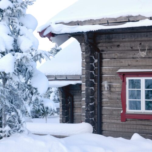 Snedækket træhus med røde vinduesrammer ved siden af snedækket gran i vinterlandskab.