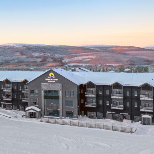 Stöten Ski Lodge med snedækkede tage, store vinduer og balkoner med udsigt til skiløjperne under en lyserød aftenhimmel.