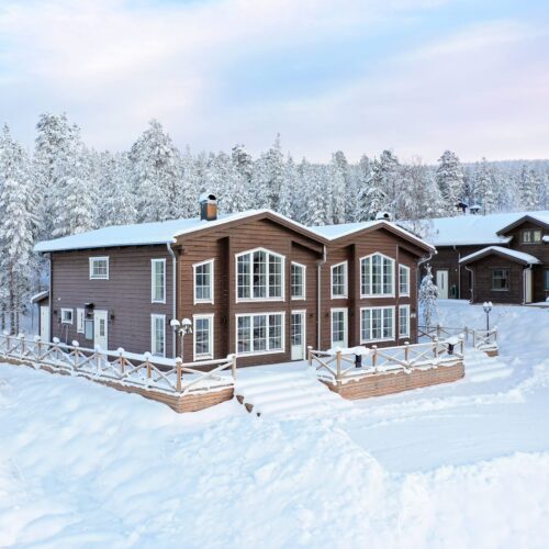 Stort tvåvåningshus i trä med stora fönster, omgiven av snötäckta träd och vinterlandskap.