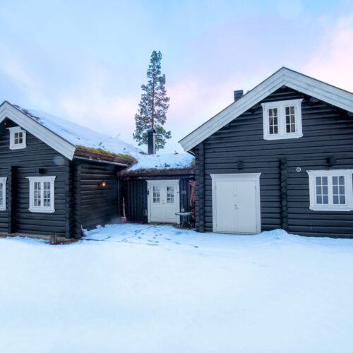 Traditionell timmerstuga med svart träpanel, vita fönster och snötäckt mark omgiven av tallar.