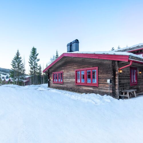 Trähus med röda detaljer i snötäckt fjällmiljö, granar och skidbacke i bakgrunden.