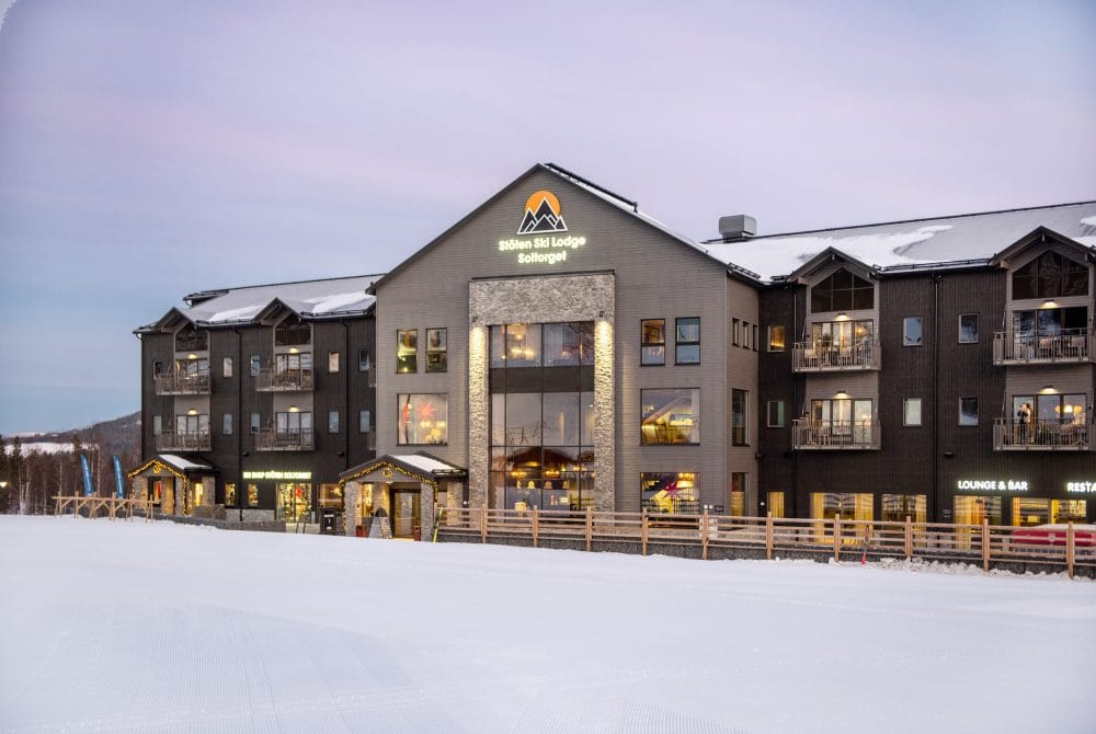 Stöten Ski Lodge med snötäckta tak, stora fönster och balkonger mot skidbackar under rosa kvällshimmel.