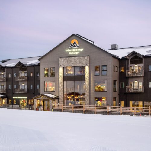 Stöten Ski Lodge med snötäckta tak, stora fönster och balkonger mot skidbackar under rosa kvällshimmel.