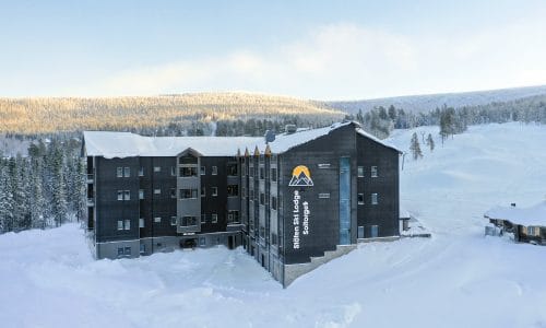 Stöten Ski Lodge med snedækkede tage, mørk træfacade og udsigt over hvide skiløjper og bjergskov.