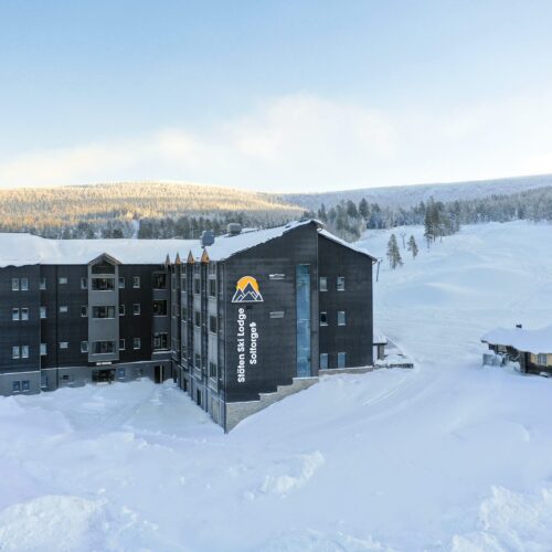 Stöten Ski Lodge med snötäckta tak, mörk träfasad och utsikt över vita skidbackar och fjällskog.