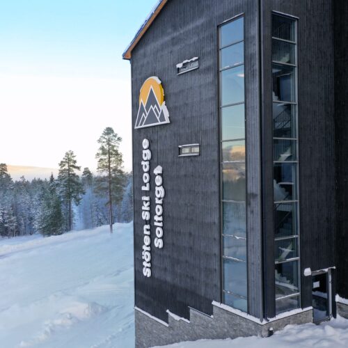 Stöten Ski Lodge fasad med svart träpanel, stora fönster och logotyp mot snötäckt fjällskog.