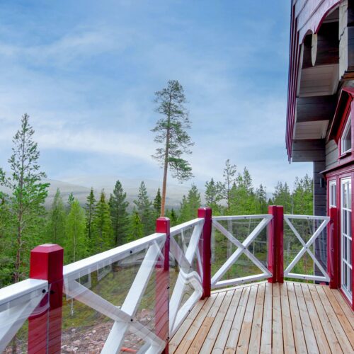 Träaltan med vit och röd räcke på fjällstuga, utsikt över grön skog och blå himmel