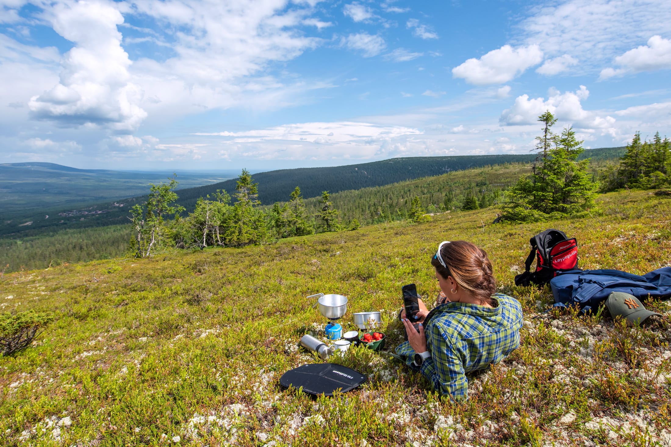 Person ligger på fjällhed med stormkök och ryggsäck, utsikt över skogsklädda berg