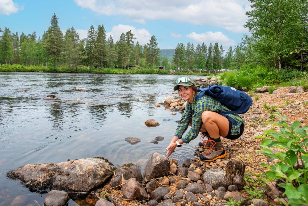 Person washing hands on rocky riverbank with rucksack, surrounded by pine forest