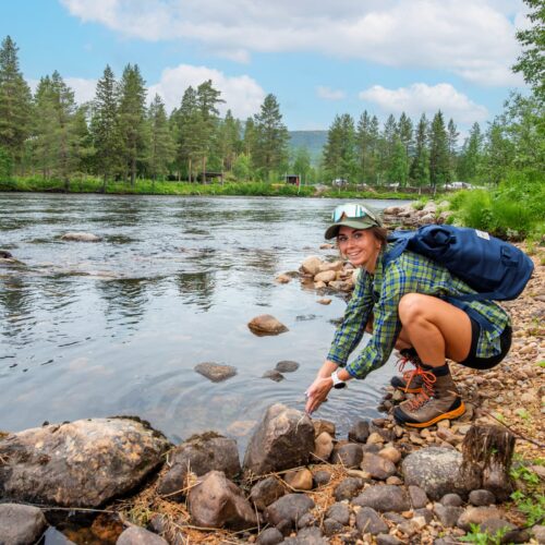 Person tvättar händerna vid stenig älvstrand med ryggsäck, omgiven av tallskog