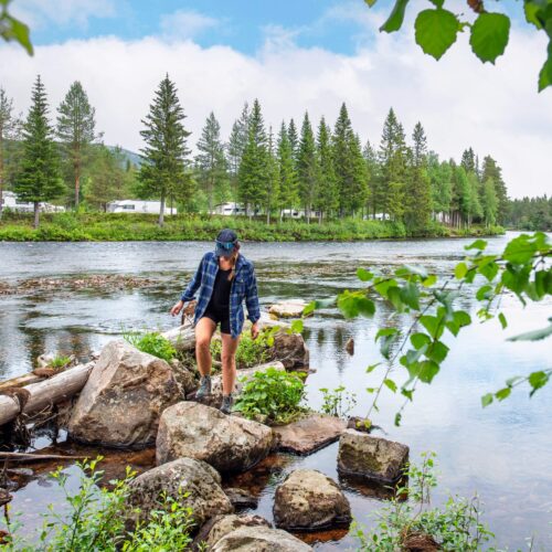 Person kliver på stenar vid lugn älv omgiven av tallskog och husvagnar