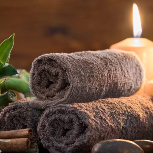 Två hoprullade handdukar med tända ljus, gröna blad, kanelstänger och skål med rosa saltskrubb.