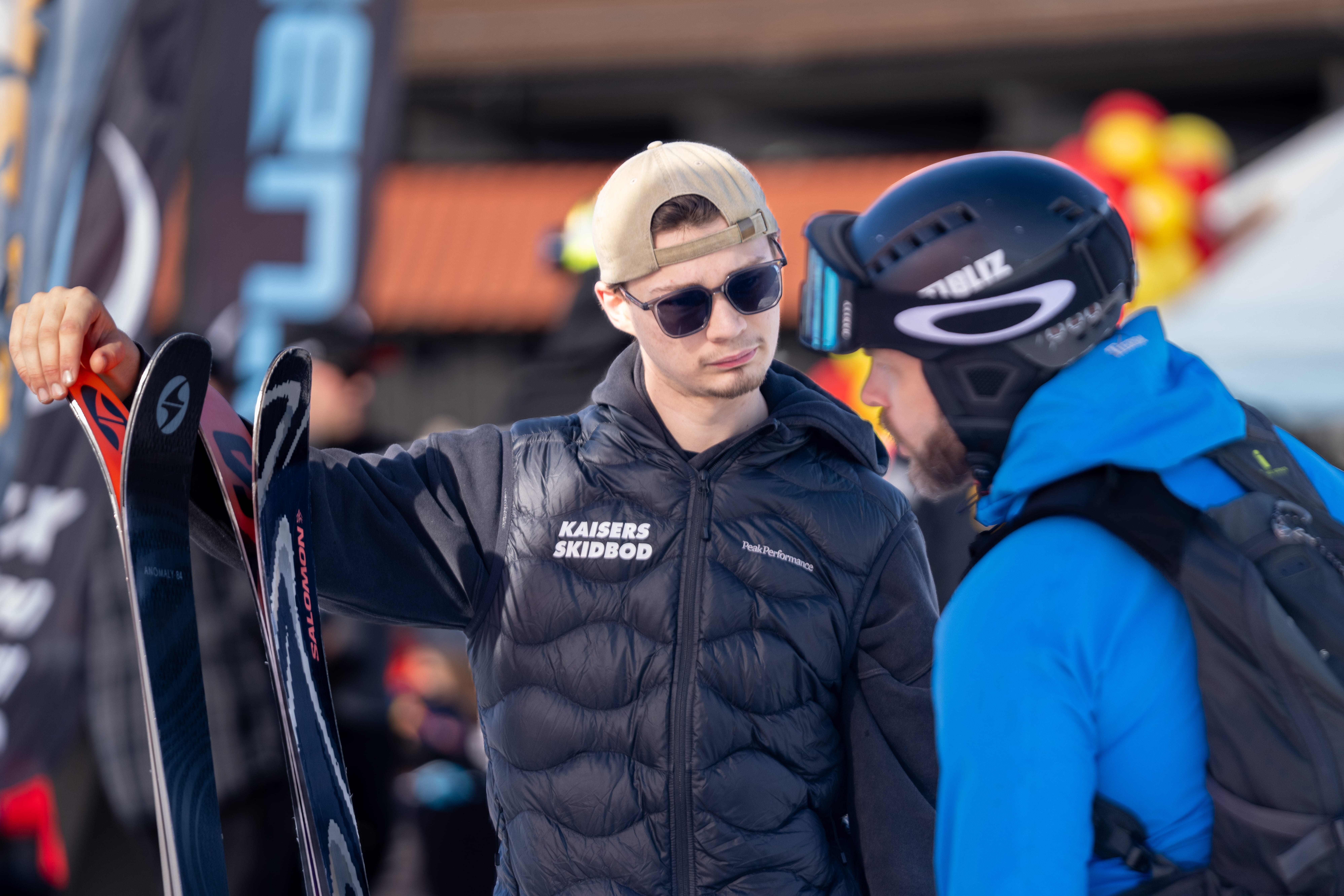 To personer i en skiudlejningsbutik diskuterer ski med hjelme, solbriller og mærkeflag i baggrunden.
