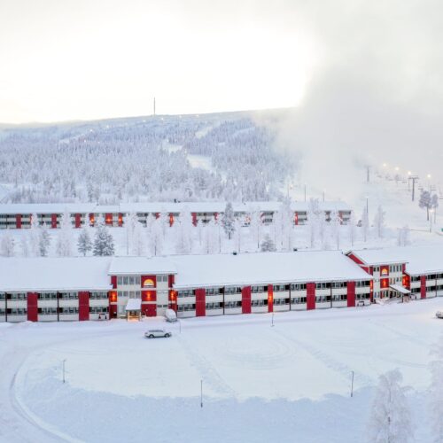 Rød og hvid bygning med flere etager, snedækket jord og parkerede biler, omgivet af frostklare træer og skiløjper med lifte i baggrunden.
